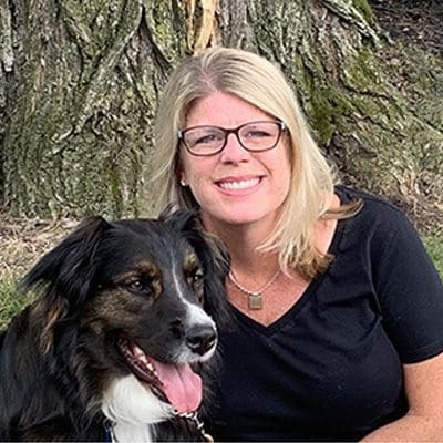 CaroandNigel A smiling blonde woman wearing glasses sits beside a black and white dog with its tongue out, in front of a tree.