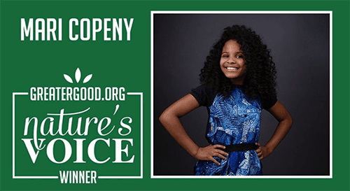 mari-c Smiling woman with curly hair wearing a blue dress, standing with hands on hips beside the GreaterGood.org Nature's Voice Winner banner.