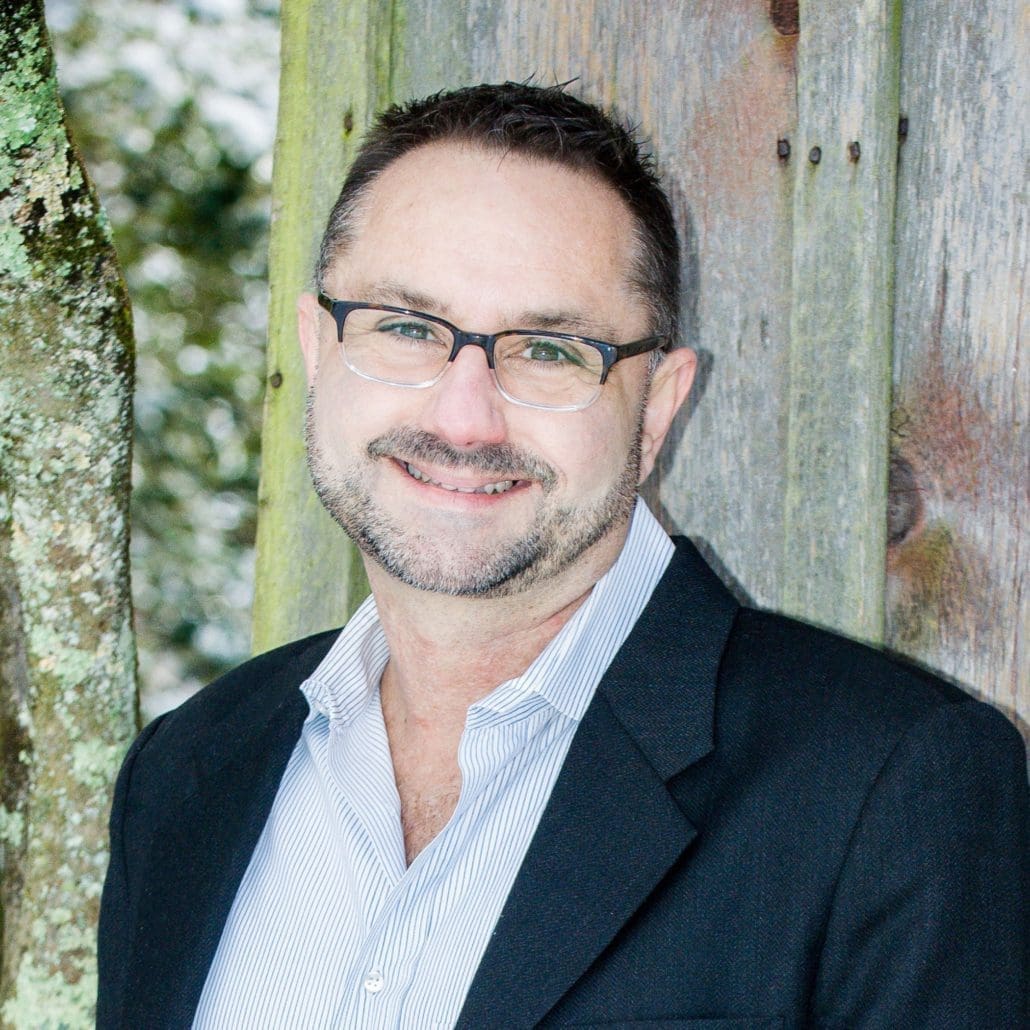 David-mugshot copy Smiling man wearing glasses, a dark blazer, and a light shirt leans against a weathered wooden fence outdoors.