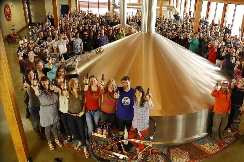 2013_new_belgium_co-workers_-_want_a_beersmall_0 Large crowd gathered around a giant copper fermentation tank in a brewery, many holding beer bottles and posing for the photo.