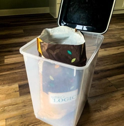 Container holding an open pet food bag sits on a wooden floor with a hinged lid open.