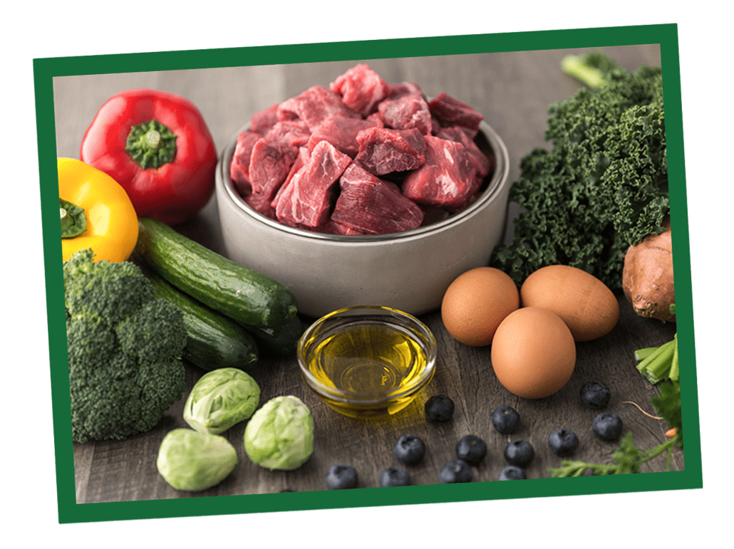 Assorted fresh ingredients on a cutting board: red bell pepper, bowl of chopped onions, garlic, Brussels sprouts, a bottle of olive oil, and eggs.