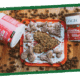 A glass dish of brown granola with a spoon on top, set on a green surface beside snack packaging.