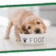 A small light-brown puppy sniffing a white bowl labeled FOOD.